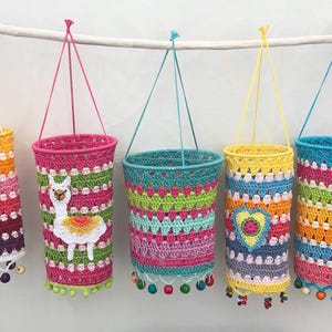 May include: Five cylindrical crochet hanging baskets in vibrant colors, suspended from a white branch. Each basket features intricate multi-colored patterns, hanging strings, and small wooden beads at the base. One basket displays a white llama applique, and another features a central heart design.