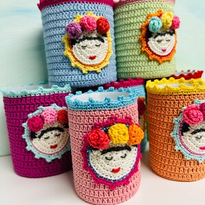 May include: A collection of cylindrical crochet covers in various colors including light blue, light green, magenta, pink, and orange. Each cover features a central crocheted face with a floral crown, closed eyes, and red lips, finished with a scalloped edge.