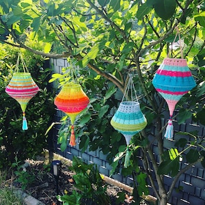 May include: Four crocheted hanging lanterns in different colors and patterns. The lanterns are hanging from a tree branch. The lanterns are made of yarn and have a decorative fringe at the bottom.
