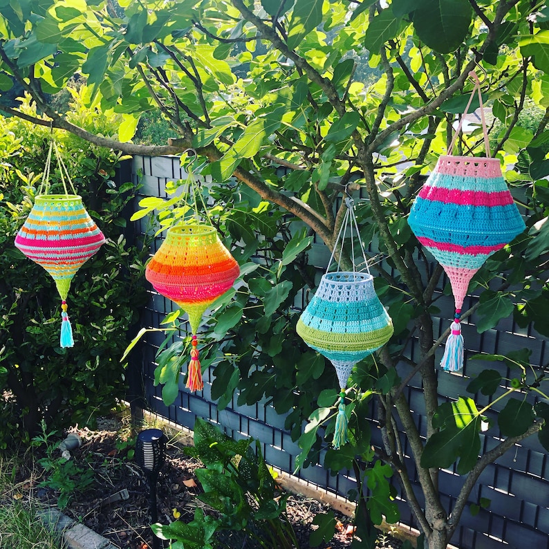 May include: Four crocheted hanging lanterns in different colors and patterns. The lanterns are hanging from a tree branch. The lanterns are made of yarn and have a decorative fringe at the bottom.