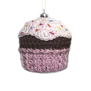 May include: A crocheted cupcake ornament featuring a light pink frosting top with multicolored yarn sprinkles, a dark brown middle section, and a light pink textured base. The ornament includes a silver-toned metal cap and a white hanging loop.