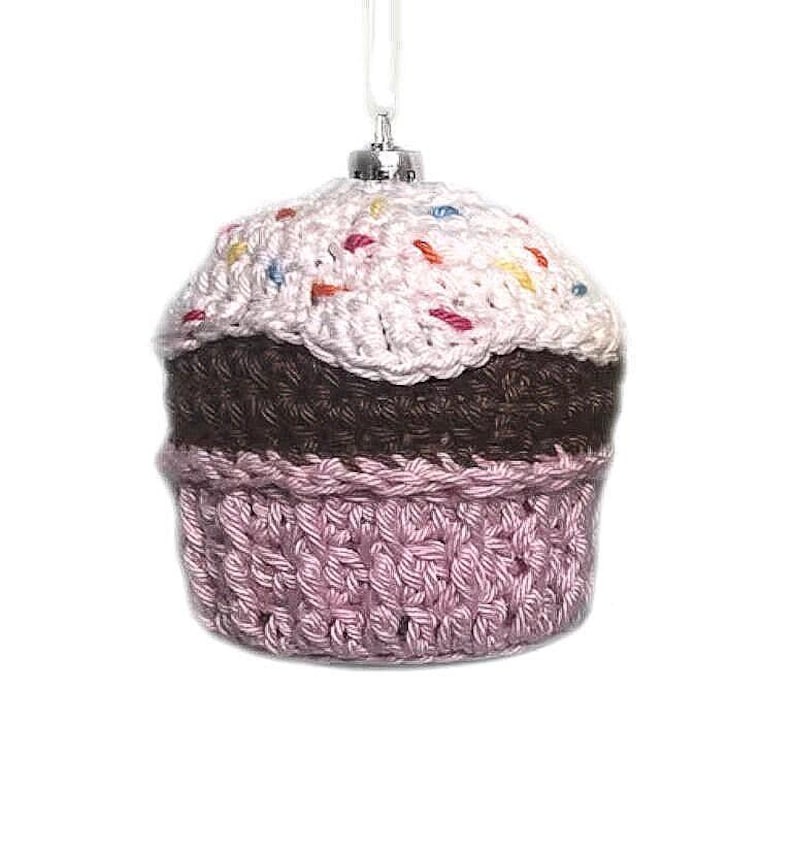 May include: A crocheted cupcake ornament featuring a light pink frosting top with multicolored yarn sprinkles, a dark brown middle section, and a light pink textured base. The ornament includes a silver-toned metal cap and a white hanging loop.