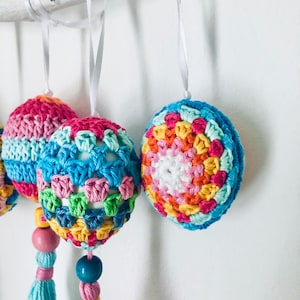K&ouml;nnte beinhalten: Ein Set aus drei bunten geh&auml;kelten Ostereiern mit Quasten, die an einem Ast h&auml;ngen. Die Eier sind in verschiedenen Farben gehalten, darunter Rosa, Blau, Gr&uuml;n, Gelb und Orange. Jedes Ei hat ein anderes Muster und ist mit einer Holzperle verziert.