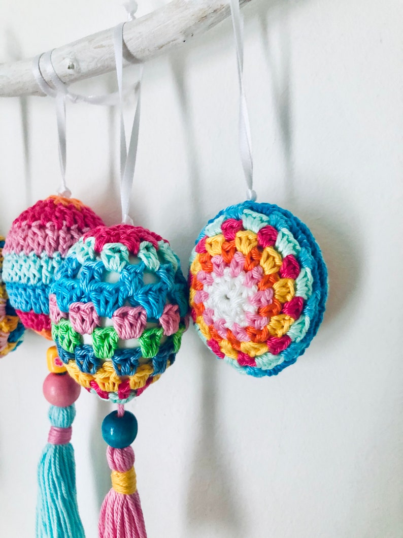 K&ouml;nnte beinhalten: Ein Set aus drei bunten geh&auml;kelten Ostereiern mit Quasten, die an einem Ast h&auml;ngen. Die Eier sind in verschiedenen Farben gehalten, darunter Rosa, Blau, Gr&uuml;n, Gelb und Orange. Jedes Ei hat ein anderes Muster und ist mit einer Holzperle verziert.