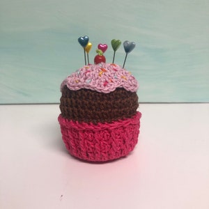 May include: A crocheted cupcake pincushion featuring a bright pink base, a dark brown middle section, and a light pink top with colorful yarn sprinkles. A small red bead sits in the center, surrounded by five pins with colorful heart-shaped heads.