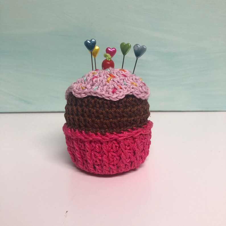 May include: A crocheted cupcake pincushion featuring a bright pink base, a dark brown middle section, and a light pink top with colorful yarn sprinkles. A small red bead sits in the center, surrounded by five pins with colorful heart-shaped heads.