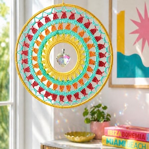 May include: A circular crochet wall hanging featuring concentric rings of yellow, orange, turquoise, and red yarn. A clear, faceted heart-shaped crystal hangs from the center of the hoop, suspended by a thin string, creating a vibrant, multi-colored design.