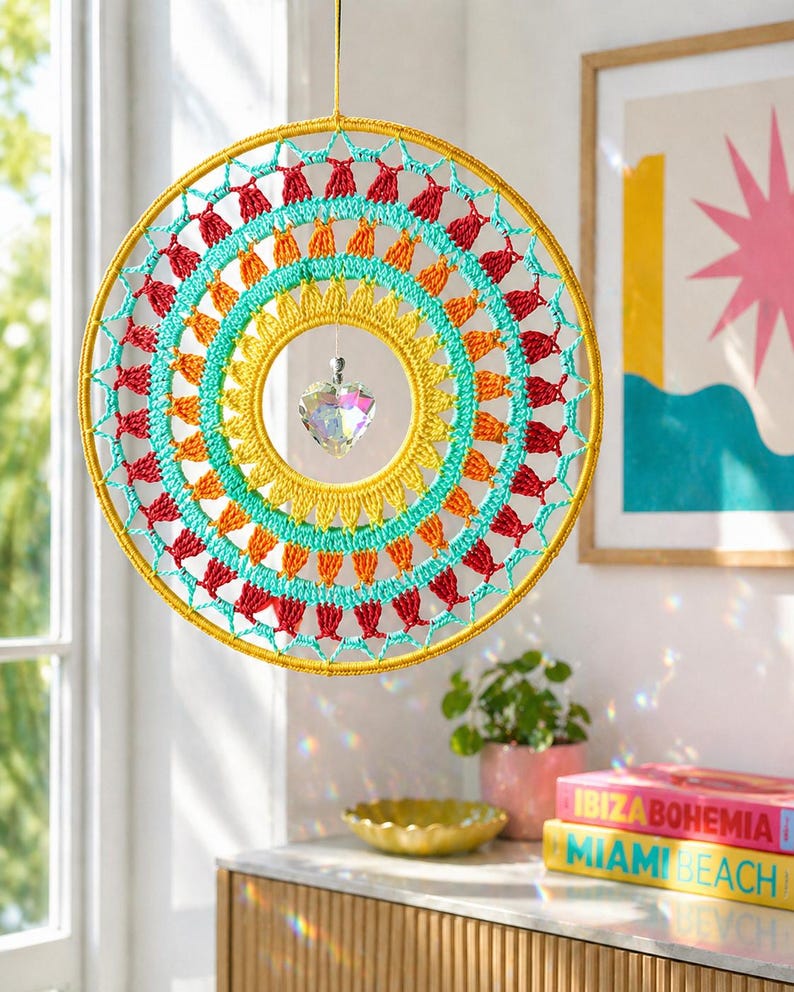 May include: A circular crochet wall hanging featuring concentric rings of yellow, orange, turquoise, and red yarn. A clear, faceted heart-shaped crystal hangs from the center of the hoop, suspended by a thin string, creating a vibrant, multi-colored design.
