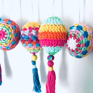 K&ouml;nnte beinhalten: Vier bunte geh&auml;kelte Ostereier mit Quasten, die an einer wei&szlig;en Schnur h&auml;ngen. Jedes Ei ist ein anderes Muster und eine andere Farbkombination, darunter rosa, blau, gr&uuml;n, gelb und orange.