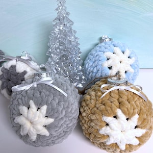 May include: A collection of handmade Christmas ornaments in various colors, including gray, blue, and tan. Each plush ornament features a white snowflake design and a silver cap with a white ribbon. A clear, decorative Christmas tree is also present.