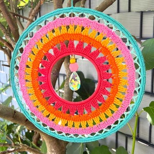 Puede incluir: Un atrapasue&ntilde;os vibrante y hecho a mano con un dise&ntilde;o de ganchillo arco&iacute;ris. El marco circular presenta anillos conc&eacute;ntricos de color rosa, naranja, amarillo y turquesa. Un colgante de cristal cuelga del centro, a&ntilde;adiendo un toque de brillo. El atrapasue&ntilde;os est&aacute; suspendido de una rama.
