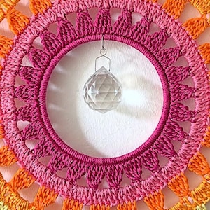 May include: A circular crochet suncatcher featuring concentric rings of orange, light pink, and dark pink yarn. A clear, multifaceted glass crystal prism hangs from a silver-toned metal loop attached to the inner edge of the central crochet ring.