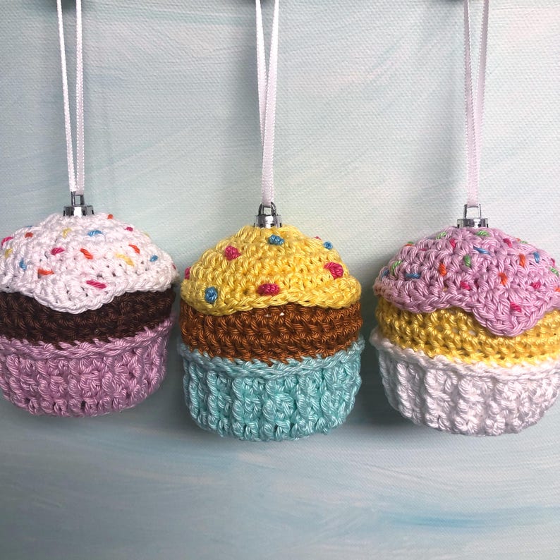 May include: Three crocheted cupcake ornaments hanging from white ribbons. The first has a white top, brown middle, and light purple base. The second has a yellow top, brown middle, and light blue base. The third has a light purple top, yellow middle, and white base.