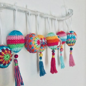 K&ouml;nnte beinhalten: Ein Set aus sieben bunten geh&auml;kelten Ornamenten mit Quasten, die an einem wei&szlig;en Ast h&auml;ngen. Die Ornamente sind aus verschiedenenfarbigen Garnen gefertigt und haben verschiedene Muster.