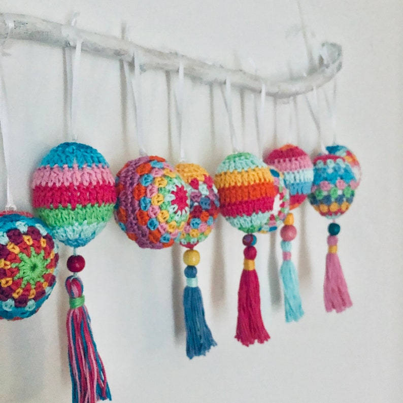 K&ouml;nnte beinhalten: Ein Set aus sieben bunten geh&auml;kelten Ornamenten mit Quasten, die an einem wei&szlig;en Ast h&auml;ngen. Die Ornamente sind aus verschiedenenfarbigen Garnen gefertigt und haben verschiedene Muster.