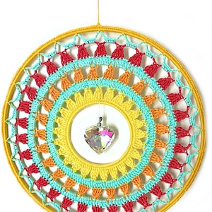 May include: A circular wall hanging featuring a yellow yarn-wrapped hoop with concentric layers of crocheted patterns in yellow, orange, turquoise, and red. A faceted, clear, heart-shaped crystal pendant hangs from the center of the hoop.