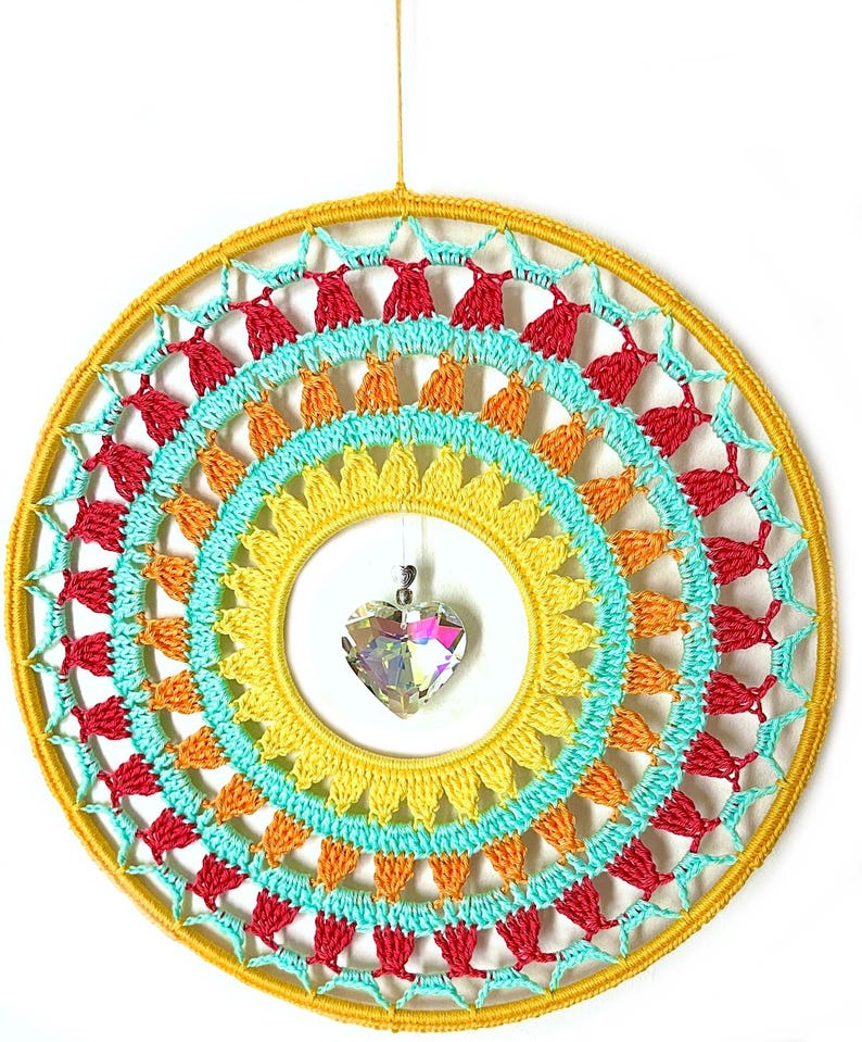 May include: A circular wall hanging featuring a yellow yarn-wrapped hoop with concentric layers of crocheted patterns in yellow, orange, turquoise, and red. A faceted, clear, heart-shaped crystal pendant hangs from the center of the hoop.