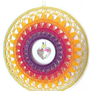 May include: A circular wall hanging featuring a crocheted mandala in concentric rings of yellow, orange, pink, and purple yarn. A clear, faceted heart-shaped crystal pendant hangs from the center of the inner purple ring by a thin thread.
