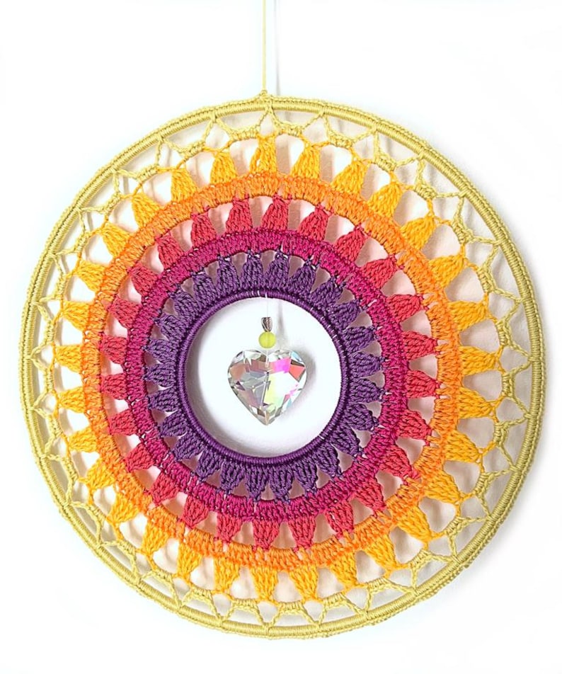 May include: A circular wall hanging featuring a crocheted mandala in concentric rings of yellow, orange, pink, and purple yarn. A clear, faceted heart-shaped crystal pendant hangs from the center of the inner purple ring by a thin thread.