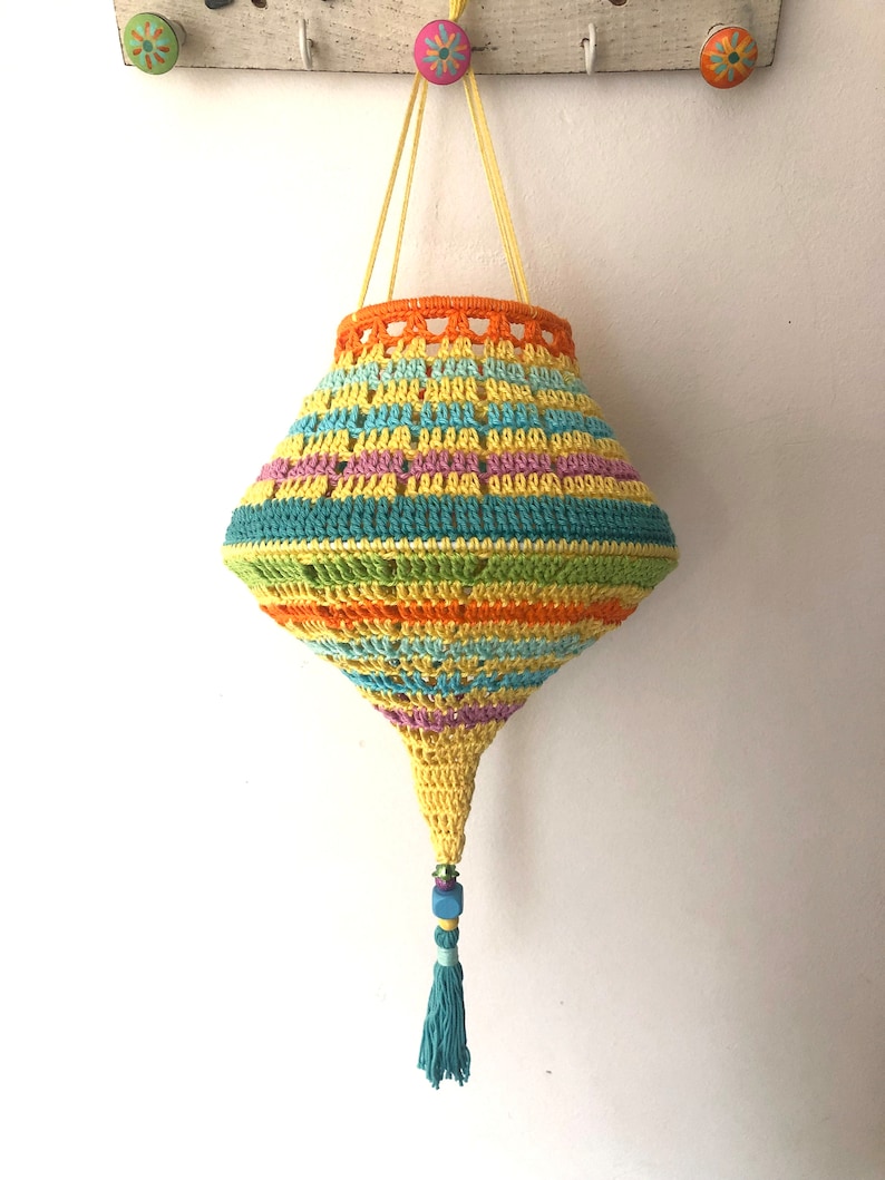 May include: A colorful crocheted hanging basket with a tassel. The basket is made with a variety of bright colors, including yellow, orange, green, blue, and purple. The basket is hanging from a white wall.