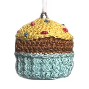 May include: A crocheted cupcake ornament featuring a pale yellow top with small pink and blue yarn accents, a brown middle section, and a light blue base. The ornament includes a silver-toned metal cap and a white ribbon loop for hanging.