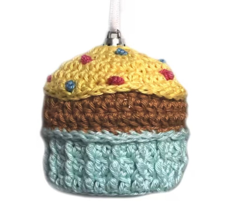 May include: A crocheted cupcake ornament featuring a pale yellow top with small pink and blue yarn accents, a brown middle section, and a light blue base. The ornament includes a silver-toned metal cap and a white ribbon loop for hanging.