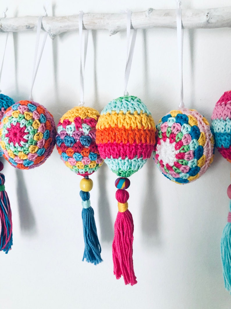 Könnte beinhalten: Ein Set aus sechs bunten gehäkelten Ostereiern mit Quasten. Jedes Ei ist eine andere Farbkombination aus Rosa, Grün, Gelb, Blau und Orange.