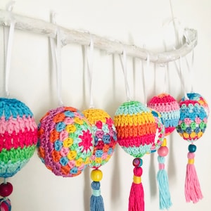 K&ouml;nnte beinhalten: Sechs bunte geh&auml;kelte Ornamente mit Quasten, die an einem wei&szlig;en Ast h&auml;ngen. Die Ornamente sind in verschiedenen leuchtenden Farben gehalten, darunter Rosa, Gr&uuml;n, Gelb, Orange und Blau. Jedes Ornament hat ein anderes Muster und ist mit einer Holzperle verziert.