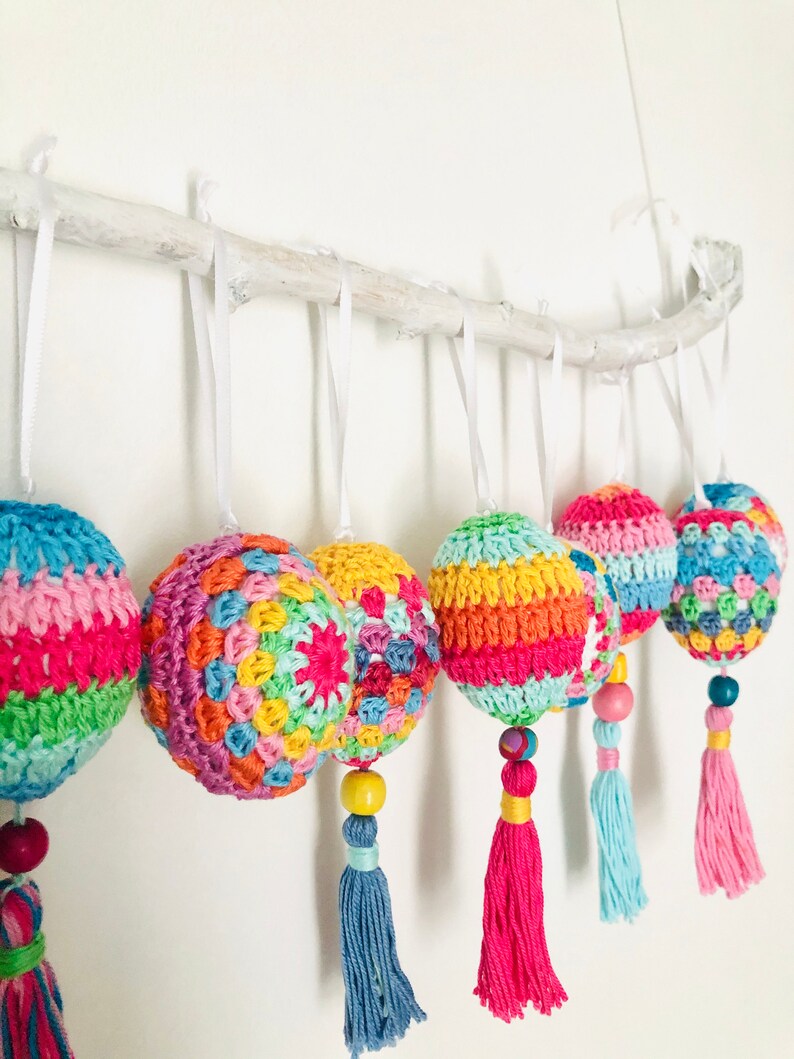 K&ouml;nnte beinhalten: Sechs bunte geh&auml;kelte Ornamente mit Quasten, die an einem wei&szlig;en Ast h&auml;ngen. Die Ornamente sind in verschiedenen leuchtenden Farben gehalten, darunter Rosa, Gr&uuml;n, Gelb, Orange und Blau. Jedes Ornament hat ein anderes Muster und ist mit einer Holzperle verziert.
