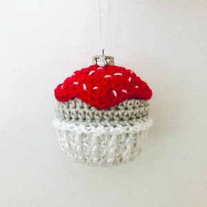 May include: A crocheted cupcake ornament featuring a bright red top with white stitched accents, a tan middle section, and a white base. The ornament is topped with a silver-toned metal cap and a thin white string for hanging purposes.