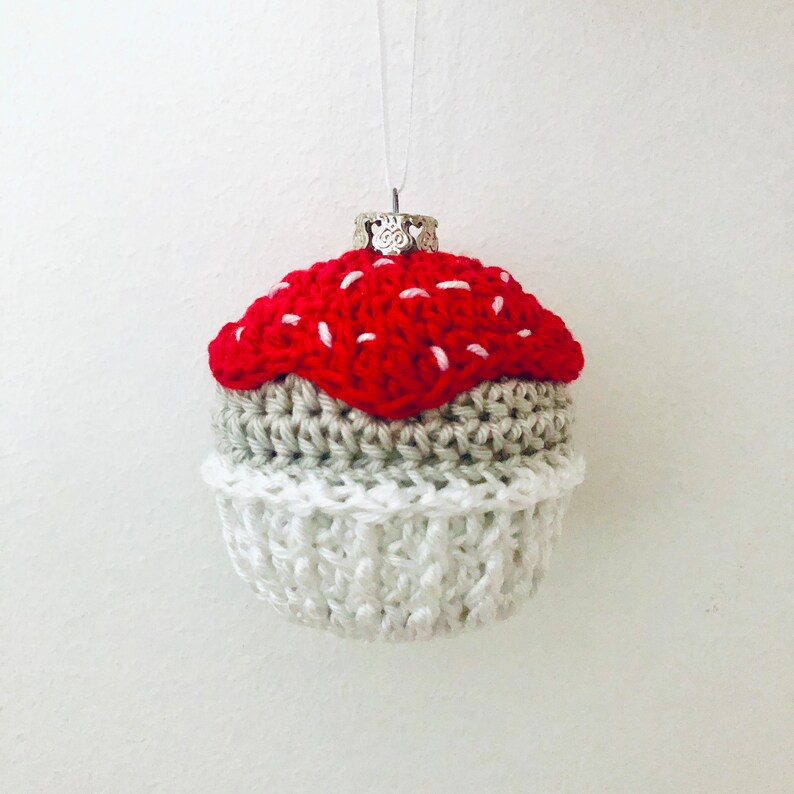May include: A crocheted cupcake ornament featuring a bright red top with white stitched accents, a tan middle section, and a white base. The ornament is topped with a silver-toned metal cap and a thin white string for hanging purposes.