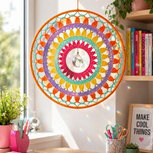 May include: A circular wall hanging featuring a vibrant, multi-colored crochet mandala pattern in orange, purple, yellow, and teal. A clear, faceted crystal prism hangs from the center of the hoop, suspended by a thin string from the top frame.