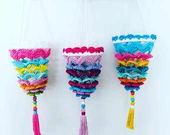 May include: Three colorful crocheted hanging planters with tassels. Each planter is made of multiple layers of crocheted fabric in different colors, including pink, blue, yellow, orange, and green. The planters are hanging from white cords attached to a white wooden surface.