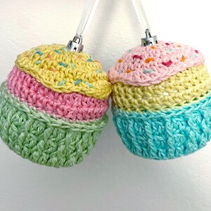 May include: Two round ornaments crocheted to resemble cupcakes, each topped with a silver cap and white ribbon. The left ornament features yellow, pink, and green yarn with colorful sprinkles. The right ornament features pink, yellow, and blue yarn.