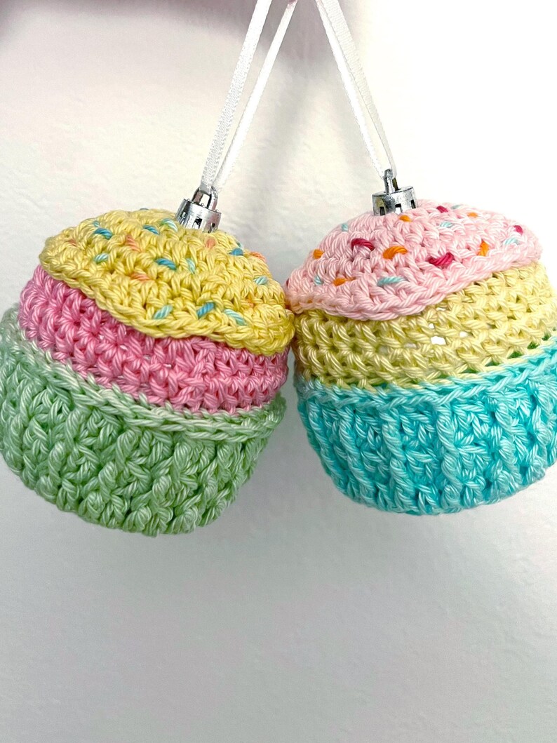 May include: Two round ornaments crocheted to resemble cupcakes, each topped with a silver cap and white ribbon. The left ornament features yellow, pink, and green yarn with colorful sprinkles. The right ornament features pink, yellow, and blue yarn.
