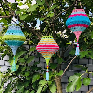 May include: Three crocheted hanging lanterns with colorful stripes and tassels. The lanterns are hanging from a tree branch in front of a gray fence.