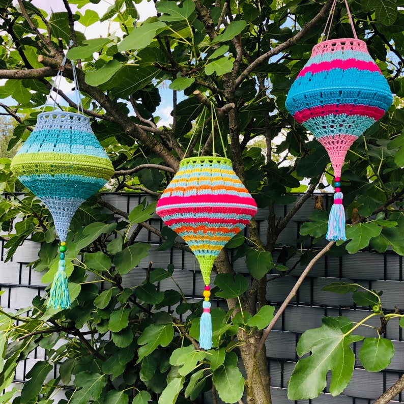 May include: Three crocheted hanging lanterns with colorful stripes and tassels. The lanterns are hanging from a tree branch in front of a gray fence.