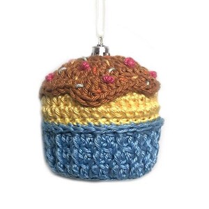 May include: A crocheted cupcake ornament featuring a blue base, a yellow middle section, and a brown top with small pink and silver accents. The ornament includes a silver-toned metal cap and a white string loop for hanging purposes.