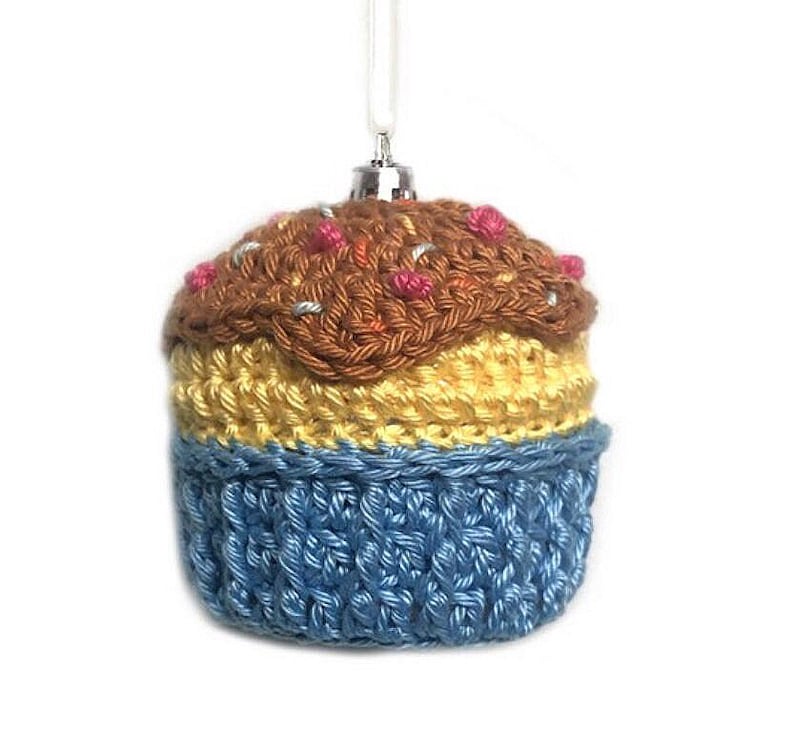 May include: A crocheted cupcake ornament featuring a blue base, a yellow middle section, and a brown top with small pink and silver accents. The ornament includes a silver-toned metal cap and a white string loop for hanging purposes.