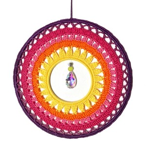 Puede incluir: Un atrapasue&ntilde;os colorido, hecho a mano, con un dise&ntilde;o de ganchillo arco&iacute;ris. El marco circular presenta tonos de rosa, naranja y amarillo, con un borde p&uacute;rpura. Un cristal en forma de l&aacute;grima cuelga del centro, a&ntilde;adiendo un toque de brillo.