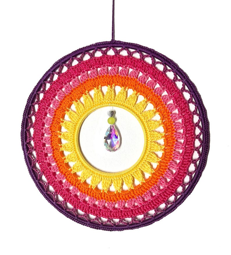 Puede incluir: Un atrapasue&ntilde;os colorido, hecho a mano, con un dise&ntilde;o de ganchillo arco&iacute;ris. El marco circular presenta tonos de rosa, naranja y amarillo, con un borde p&uacute;rpura. Un cristal en forma de l&aacute;grima cuelga del centro, a&ntilde;adiendo un toque de brillo.