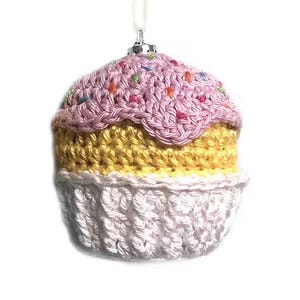 May include: A crocheted cupcake ornament featuring a white base, a yellow middle section, and a pink top decorated with small, multi-colored yarn sprinkles. The ornament includes a silver-toned metal cap and a white hanging loop at the top.