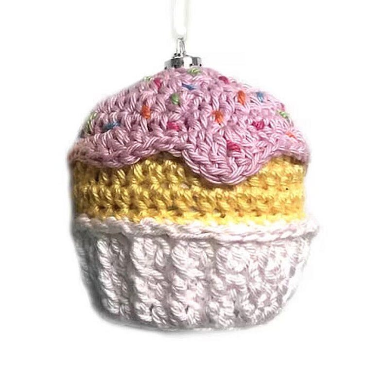 May include: A crocheted cupcake ornament featuring a white base, a yellow middle section, and a pink top decorated with small, multi-colored yarn sprinkles. The ornament includes a silver-toned metal cap and a white hanging loop at the top.