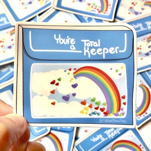 May include: A blue and white sticker with a rainbow and hearts. The sticker says "You're a Total Keeper" and has the artist's social media handle at the bottom.