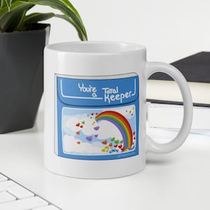 May include: White ceramic mug with a blue design featuring a rainbow, hearts, and clouds. The mug has the text "You're a Total Keeper" in a handwritten style. The mug is on a white desk with a keyboard and a plant in the background.