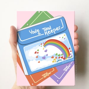May include: A blue binder-style greeting card featuring the text "You're a Total Keeper" above a graphic of a rainbow and colorful hearts floating among white clouds. The design includes smaller orange and purple binder accents in the background.