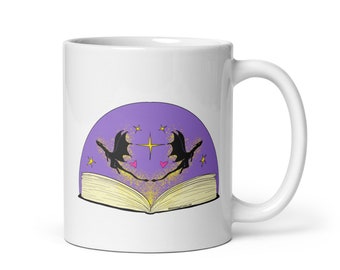May include: White ceramic mug featuring a fantasy illustration. The design includes a purple arch with two black dragon silhouettes, stars, and hearts above an open book. The mug has a white handle and a glossy finish.