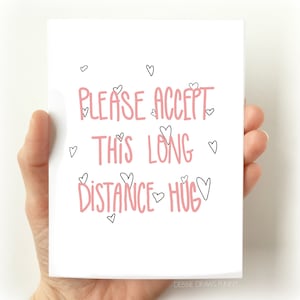 May include: A white card with pink text that reads "Please accept this long distance hug" surrounded by small black hearts. The card is being held by a hand.