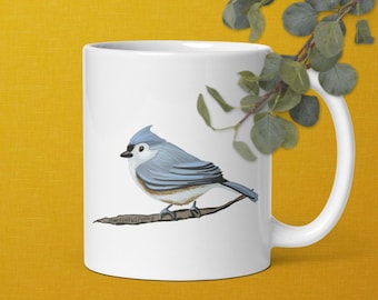 Puede incluir: Taza de cerámica blanca con una ilustración de un pájaro azul y gris posado en una rama. El pájaro tiene una cresta azul y una barriga blanca. La taza tiene un borde y un asa negros.