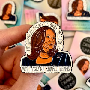Op de afbeelding: Een witte emaille pin met een portret van Kamala Harris, vice-president van de Verenigde Staten. De pin heeft de tekst "The American Dream Belongs To All Of Us" en "Vice President Kamala Harris".